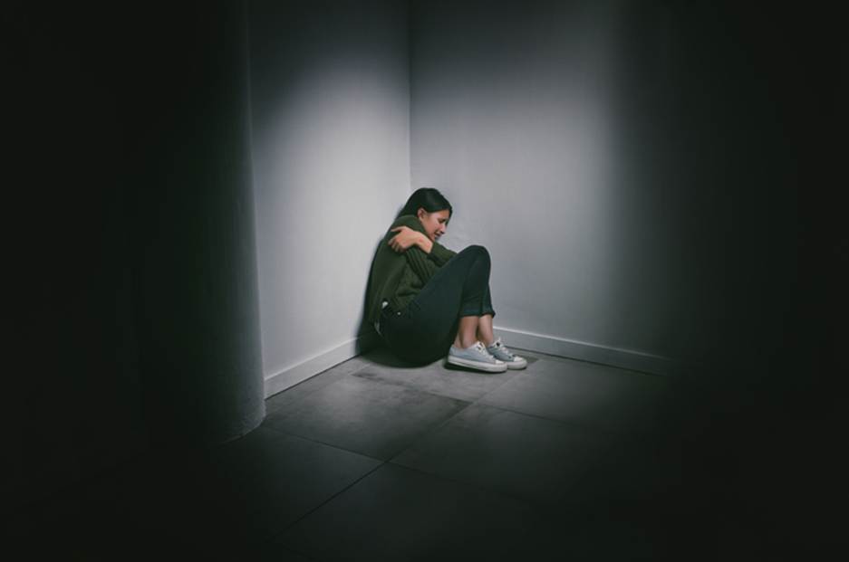 A person crouches in a dark corner, symbolizing the hidden nature of abuse discussed in the article about misconduct in healthcare settings. The image aims to highlight the emotional impact on victims.